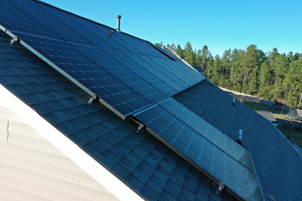 Drone view of a flush-mounted solar panel array on a residential shingle roof, surrounded by mature trees, South Carolina Midlands.